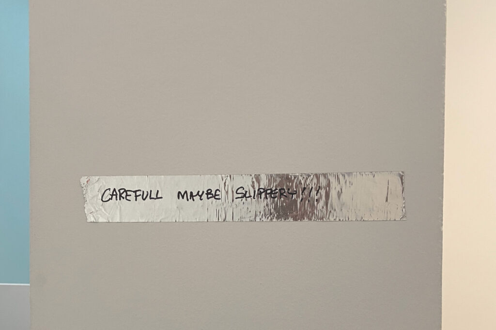 Devon Dikeou, Tape, from The Inconspicuouses at MCA Denver — a strip of silver tape on a gallery wall hand-lettered "CAREFULL MAYBE SLIPPERY!!!"